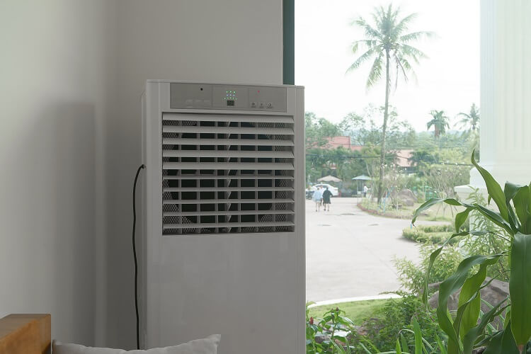 Ar-condicionado portátil colocado perto de uma janela com vista para várias folhagens. 