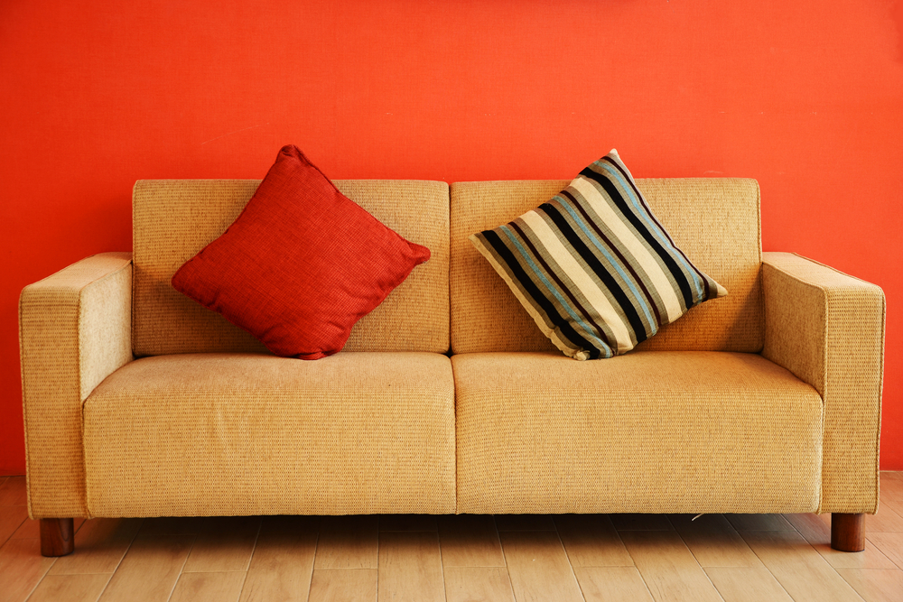 Sofá de cor neutra, com duas almofadas, colocado na frente de uma parede na cor laranja.