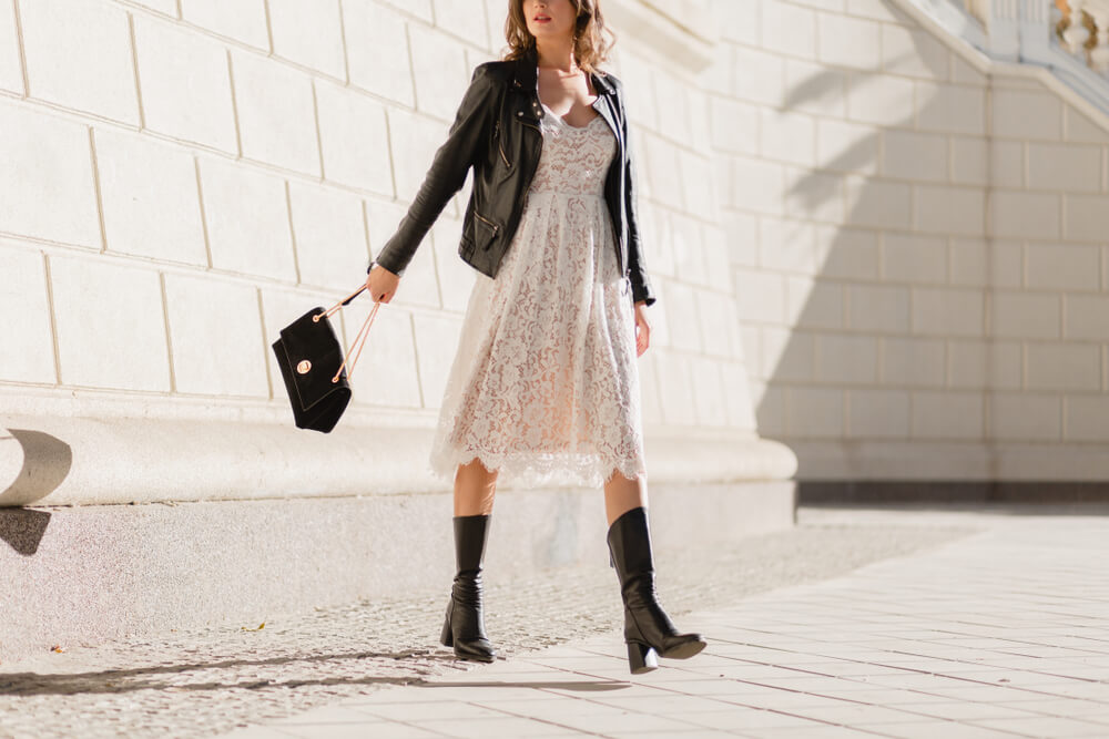 Na imagem vemos uma mulher utilizando vestido, bota e jaqueta de couro. Ela está segurando uma bolsa preta e está no meio de uma rua.