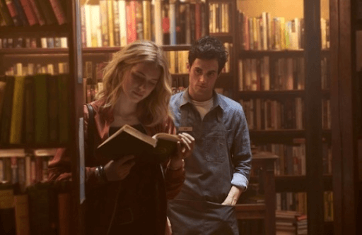 Na imagem vemos um homem e uma mulher em uma livraria. Ele observa ela a distância, que está lendo um livro.