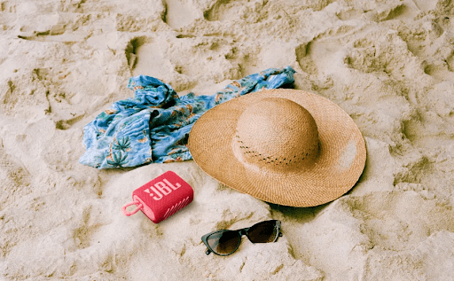 Foto de um chapéu, um óculos, uma canga e uma caixa de som JBL em uma areia.