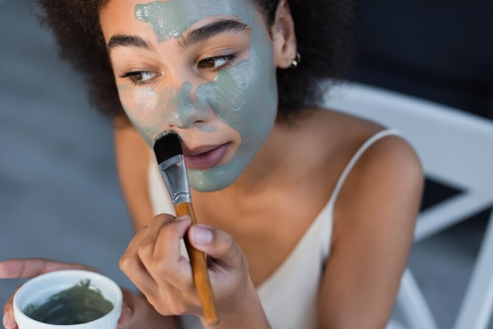 Imagem de uma mulher negra passando argila verde no rosto.