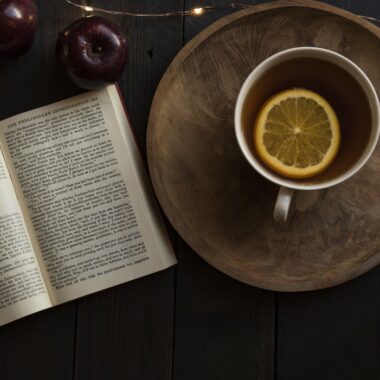 Livro aberto com xícara de chá ao lado e maçãs acima.
