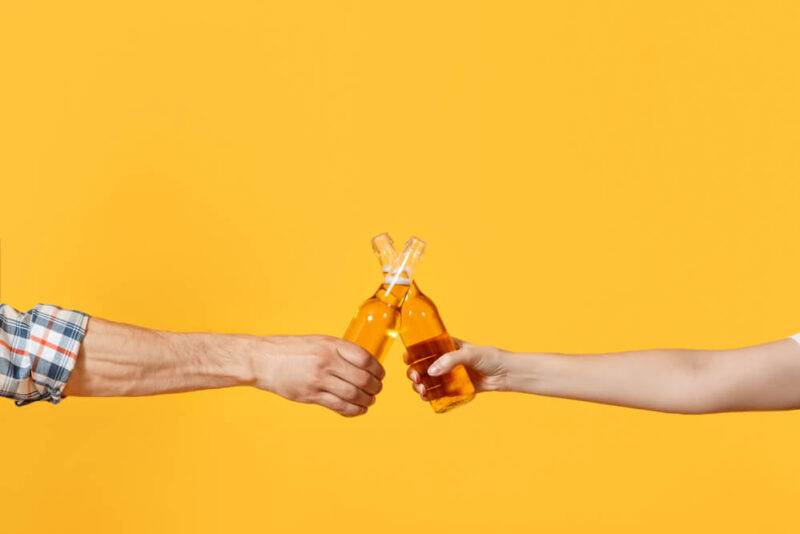 Dois braços segurando cada um uma garrafa de cerveja, em um fundo amarelo.