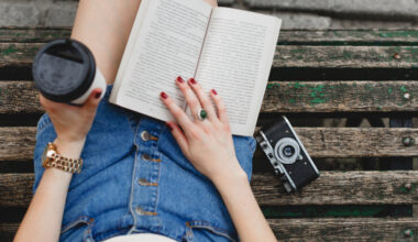 Menina sentada em um banco, usando uma saia jeans, lendo um livro, com uma câmera ao lado e um copo de café em uma das mãos.