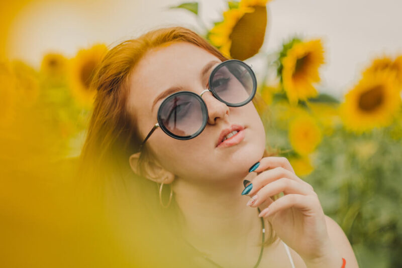 Mulher entre uma plantação de girassois utilizando um óculos diferente de sol.