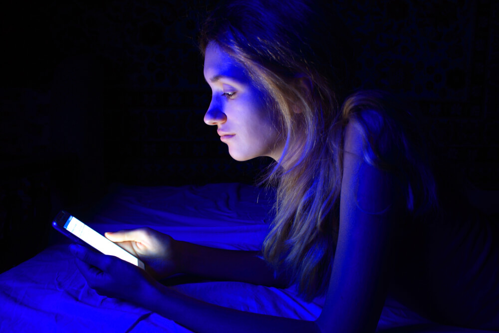 Mulher jovem deitada em uma cama, em um quarto escuro, com a luz do celular refletindo na face dela.