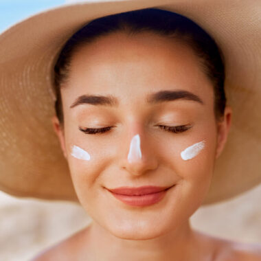 Mulher branca em uma praia, usando um chapéu e com protetor solar facial no rosto, com riscos do creme nas bochechadas e no nariz.