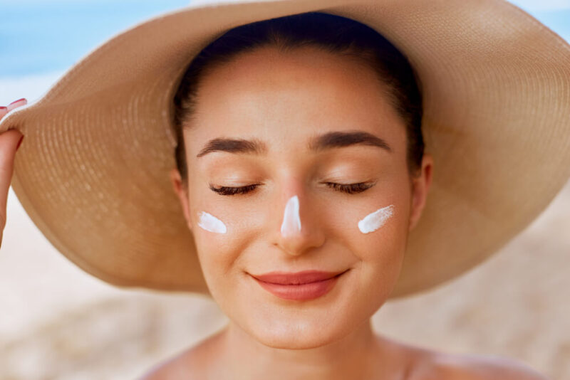 Mulher branca em uma praia, usando um chapéu e com protetor solar facial no rosto, com riscos do creme nas bochechadas e no nariz.