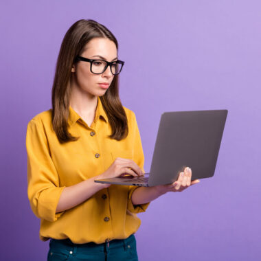 Mulher branca vestindo camiseta laranja, de óculos, enquanto segura um notebook Lenovo IdeaPad S145. Ao fundo, há uma parede roxa.