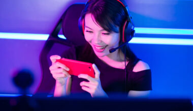 Mulher sentada em uma cadeira gamer, enquanto mexe no celular e está com um fones de ouvido gamers hyperx.