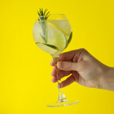 Uma mão segurando uma taça de gin tônica com limão e alecrim, em um fundo amarelo.