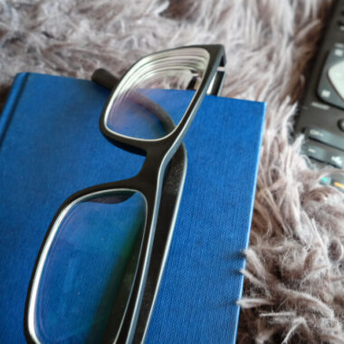 Na imagem vemos um livro de capaz azul. Em cima dele está um óculos de grau. Ao lado, há o controle de uma TV.