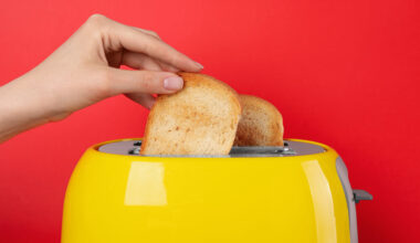 Imagem de uma torradeira amarela. Uma mão está colocando um pão nela. Ao fundo, vemos uma parede vermelha.