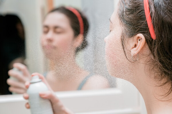 mulher de cabelos castanhos em frente a um espelho aplicando água termal no rosto.