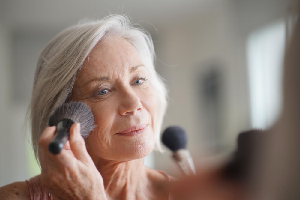 Uma mulher de meia idade, com os cabelos brancos, em frente ao espelho - se maquiando.
