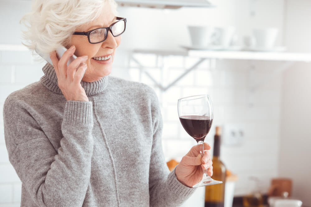Mulher de meia idade, falando no telefone, enquanto toma uma taça de vinho. Ela está sorrindo.