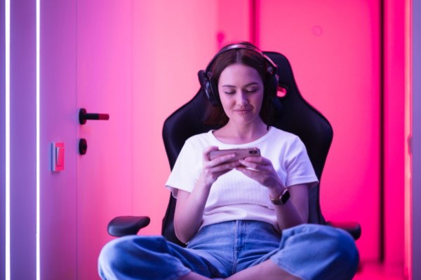 Mulher jovem branca de camiseta branca e calça jeans, sentada em cadeira gamer, usando headset como fono de ouvido gamer para celular.