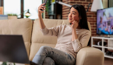 Uma mulher sentada no sofá utilizando o celular para fazer selfie.