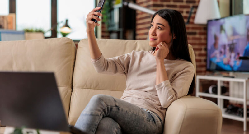 Uma mulher sentada no sofá utilizando o celular para fazer selfie.