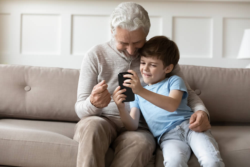Imagem de um homem idoso ao lado de uma criança. Ela está segurando um celular positivo.