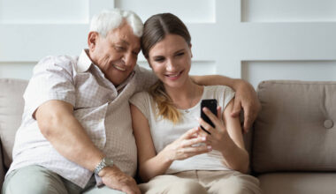 Imagem de um homem idoso ao lado de uma mulher jovem. Ela está ensinando ele a utilizar um celular positivo.