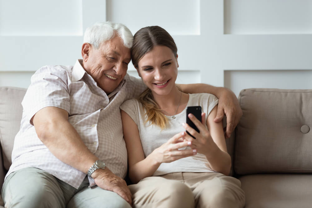 Imagem de um homem idoso ao lado de uma mulher jovem. Ela está ensinando ele a utilizar um celular positivo.