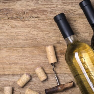 Imagem mostra duas garrafas de vinho deitadas sob uma mesa de madeira, com saca-rolhas e rolhas espalhadas pela superfície.